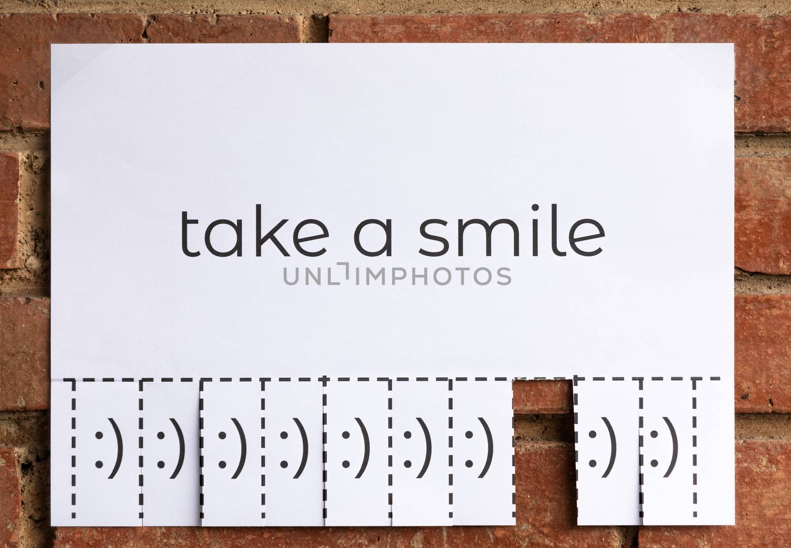 A paper with the phrase "Take a Smile" writen on it with different pieces with smile faces to tear off. Smiling is free. The image is from the front.