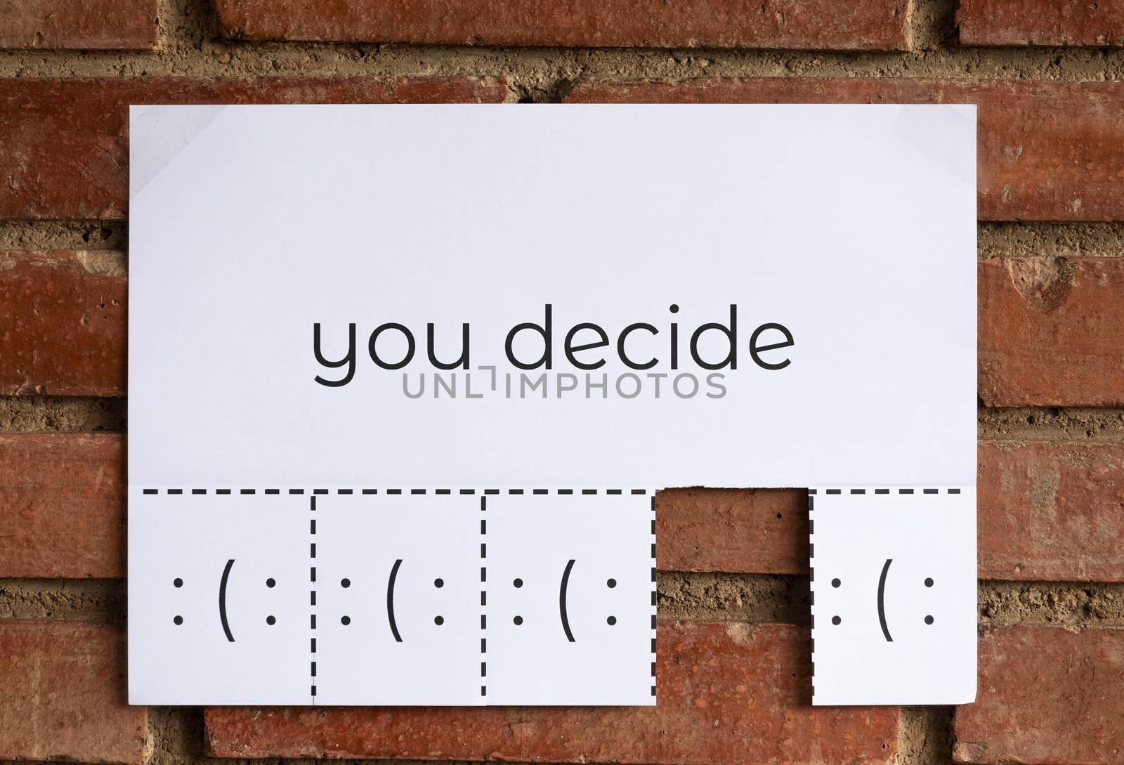 A paper with the phrase "You Decide" writen on it with different pieces with smile faces to tear off and decide what do you want to be, sad or happy. Smiling is free. The image is from the front.