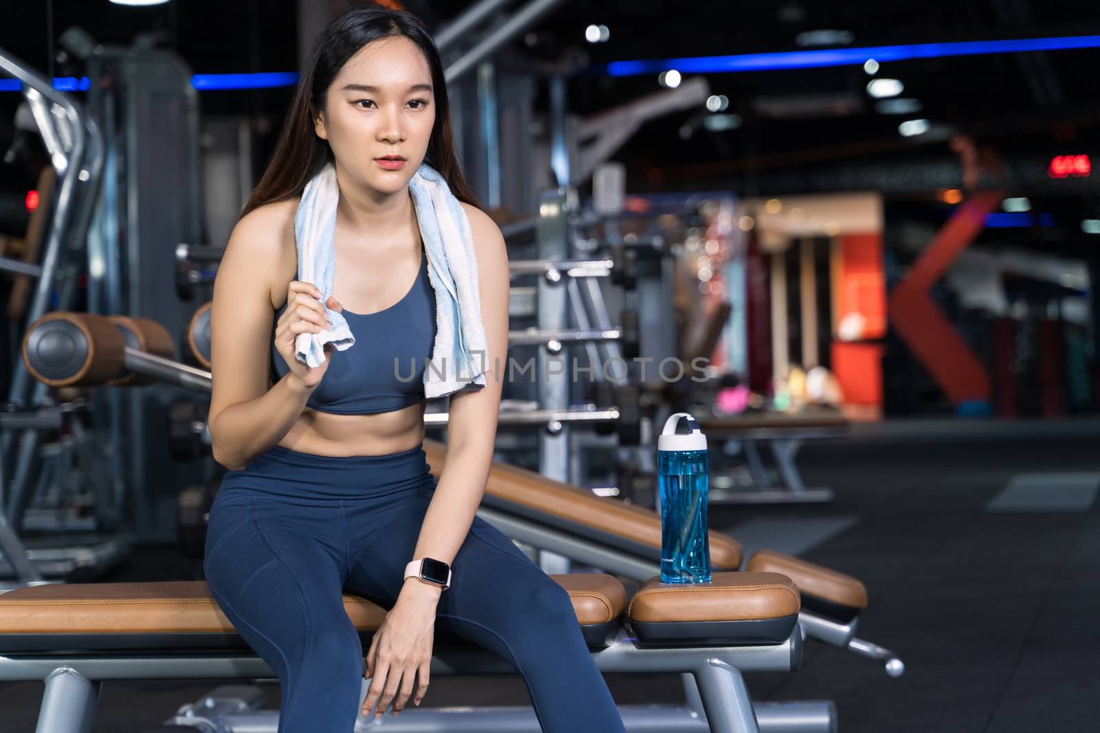 Asian women are sitting on the exercise and are holding towels with water bottles placed on their sides. She's sitting alone at the gym in sport healthy concept