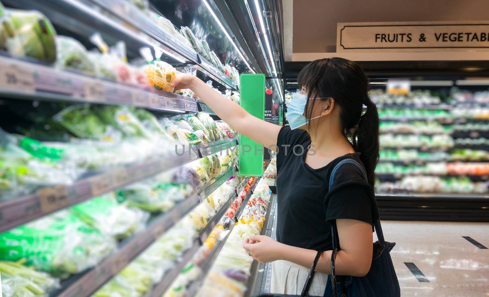 Asian women are shopping at the grocery store, holding baskets and wearing a health mask to prevent infection. She's picking vegetables from the shelves in lifestyle shopping concept