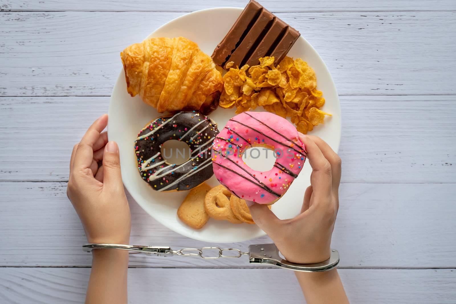 Calories control, food diet and weight loss concept. top view of two hands was control by handcuff, holding doughnut from white dish on wooden table by asiandelight