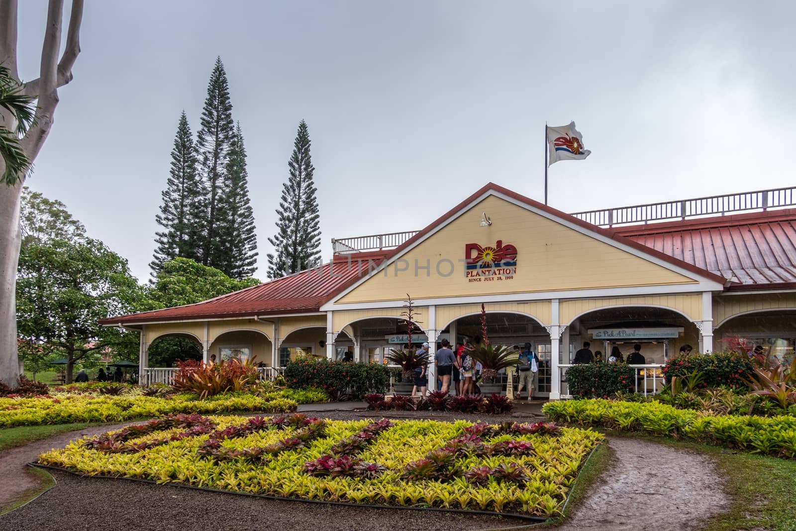 Wahiawa, Oahu, Hawaii, USA. - January 09, 2020: Welcome building, entrance to Dole pineapple plantation, museum and shop. Yellow wall, red roof, green, red and yellow vegetation under light blue sky.
