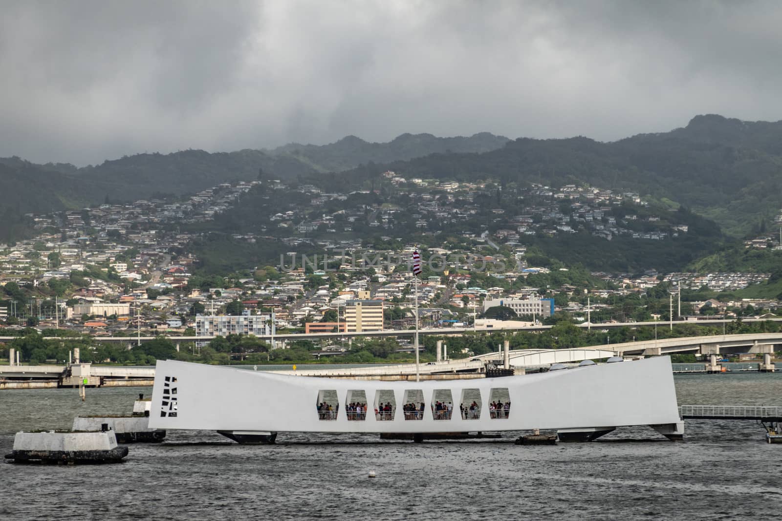 Oahu, Hawaii, USA. - January 10, 2020: Pearl Harbor. White USS Arizona Memorial and Ford Island bridge in back. Green Hills with white buildings under rainy dark cloudscape behind.
