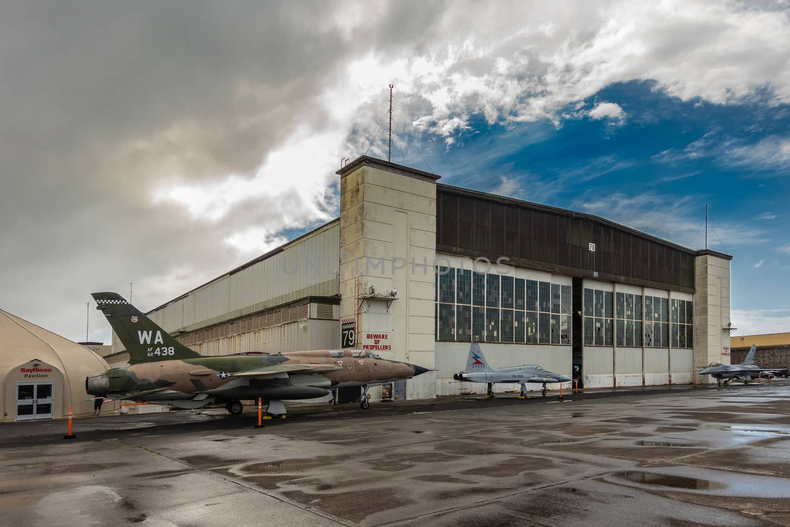 Oahu, Hawaii, USA. - January 10, 2020: Pearl Harbor Aviation Museum. 3 fighter jets outside hangar 79 on wet tarmac under rainy sky with blue patch. Raytheon pavillion nearby.