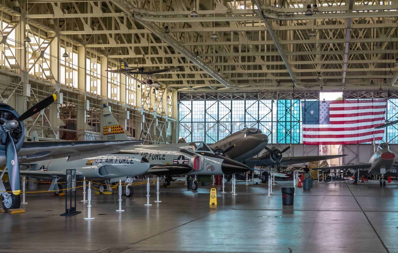 Oahu, Hawaii, USA. - January 10, 2020: Pearl Harbor Aviation Museum. Line of airplanes in hangar with large USA flag in back. Beige roof. Natural light through windows.
