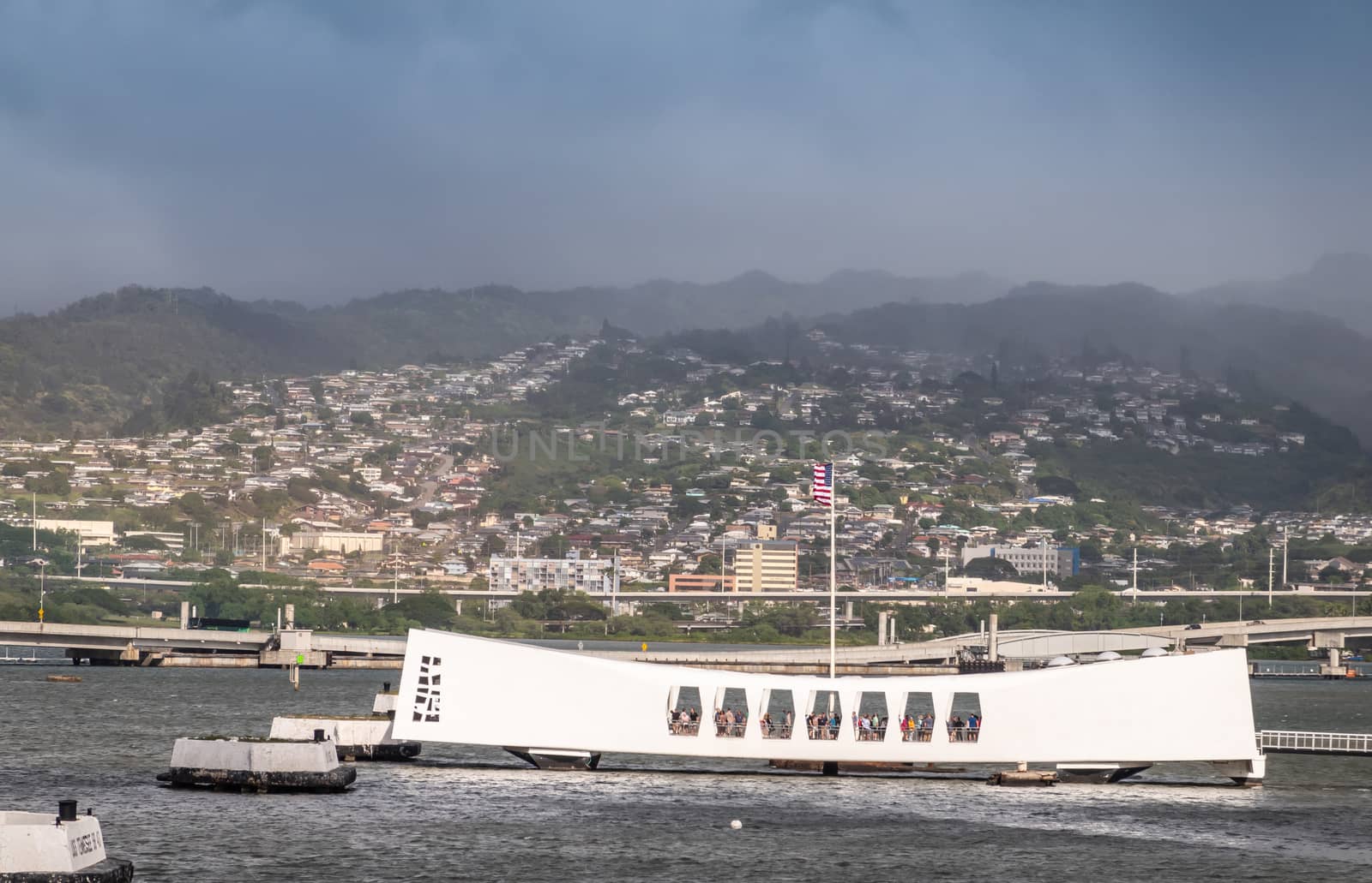 Oahu, Hawaii, USA. - January 10, 2020: Pearl Harbor. White USS Arizona Memorial and Ford Island bridge in back. Green Hills with white buildings under rainy blue cloudscape behind.