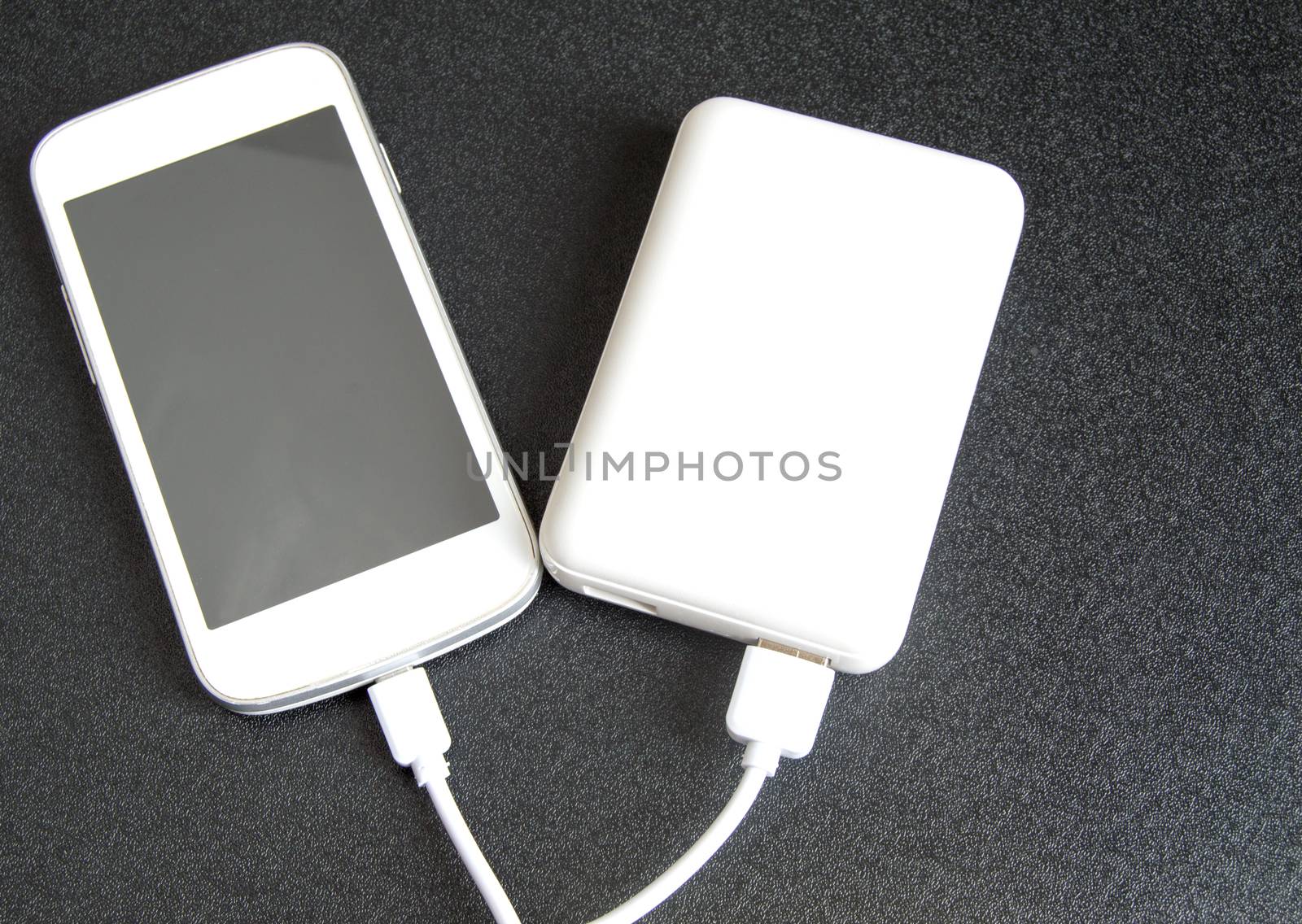 A white phone with a black screen and a copy of space is charged using a white power Bank. Top view, black background by claire_lucia