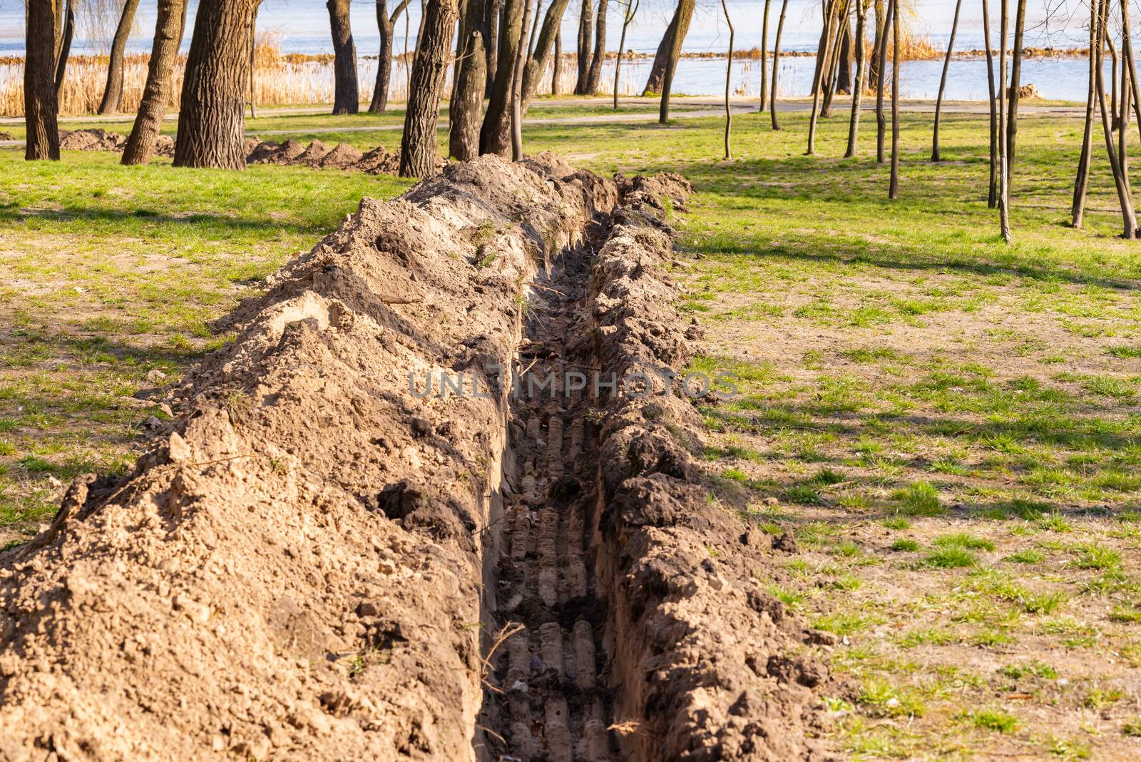A trench in the ground for laying a pipe, or tubes, in the park, under the trees, close to the river, with spring light