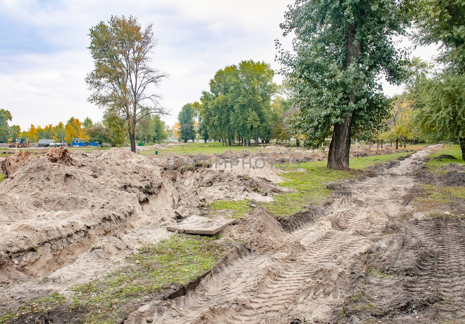 A trench in the sandy ground for laying a pipe in the park, under the trees, with evening autumn light