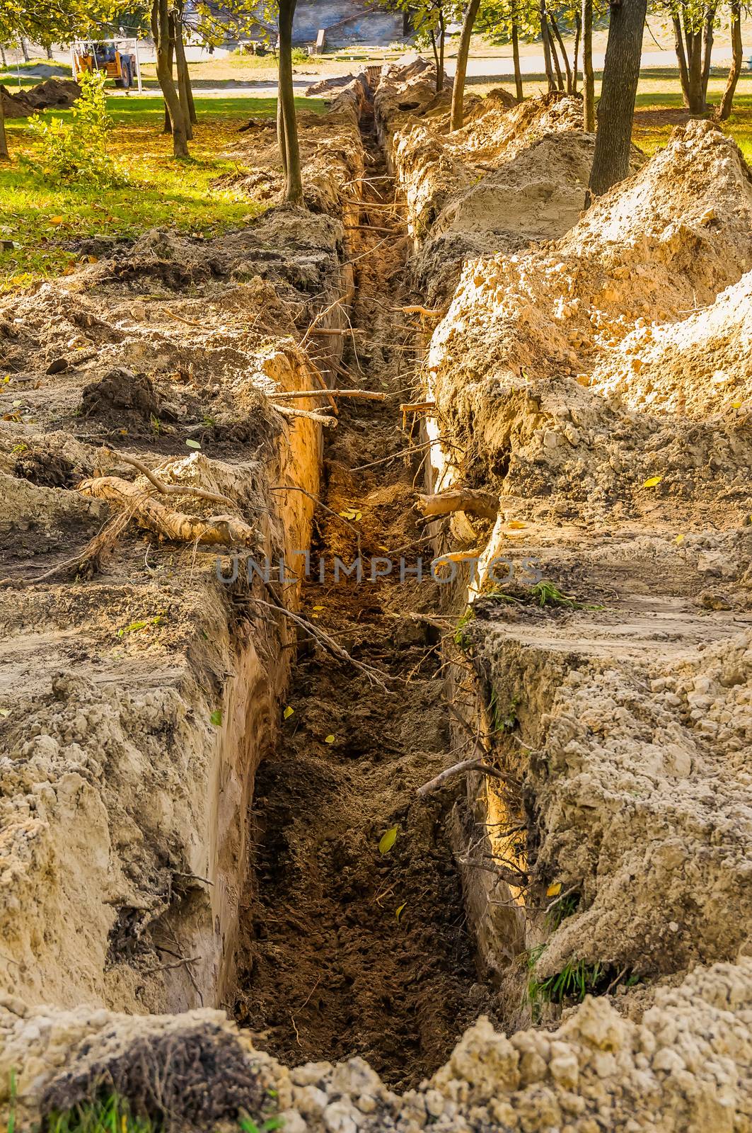 A trench in the ground for laying a pipe in the park, under the trees, with evening autumn light