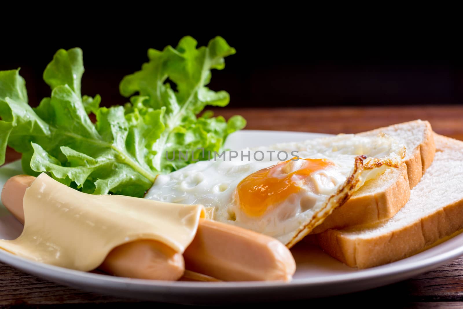 American breakfast concept. Fried egg with sliced bread, boiled chicken sausage, cheddar cheese, and fresh lettuce on a white plate in the home kitchen. Homemade food in the morning.