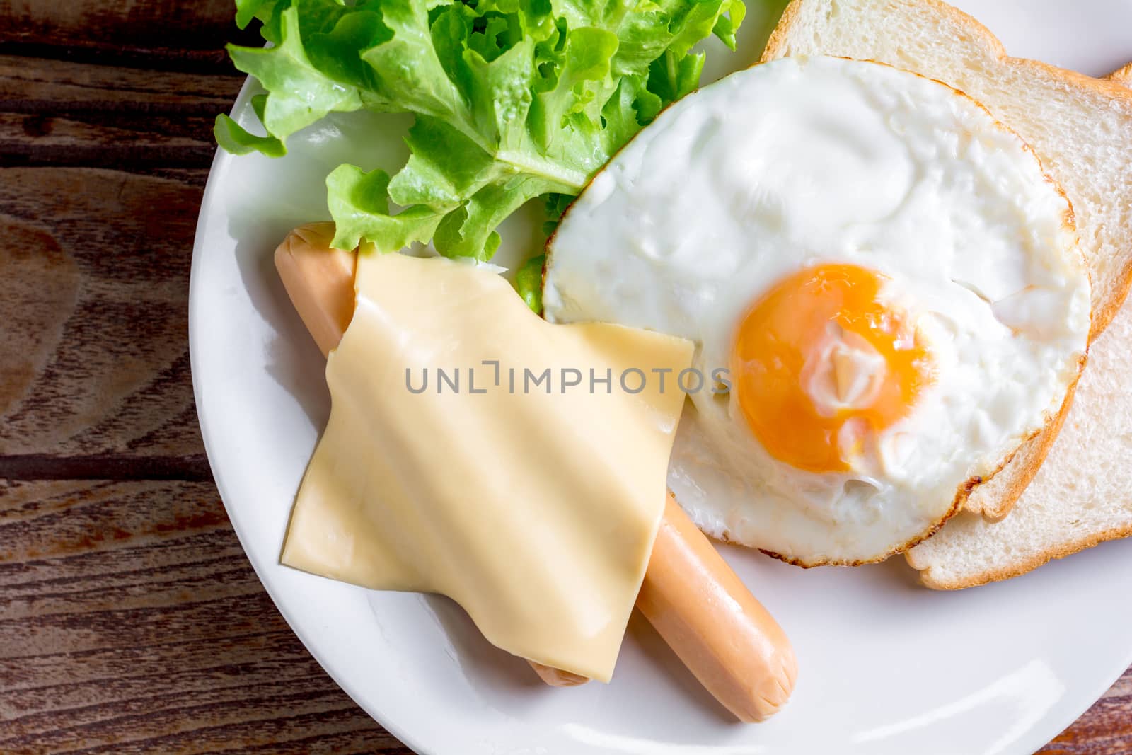 American breakfast concept. Fried egg with sliced bread, boiled chicken sausage, cheddar cheese, and fresh lettuce on a white plate in the home kitchen. Homemade food in the morning.