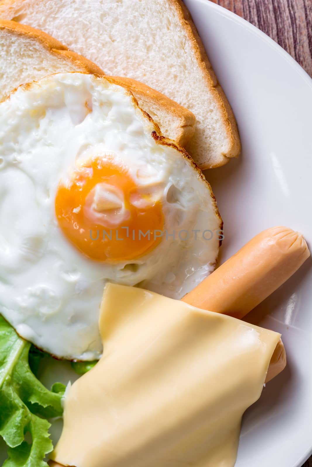 American breakfast concept. Fried egg with sliced bread, boiled chicken sausage, cheddar cheese, and fresh lettuce on a white plate in the home kitchen. Homemade food in the morning.