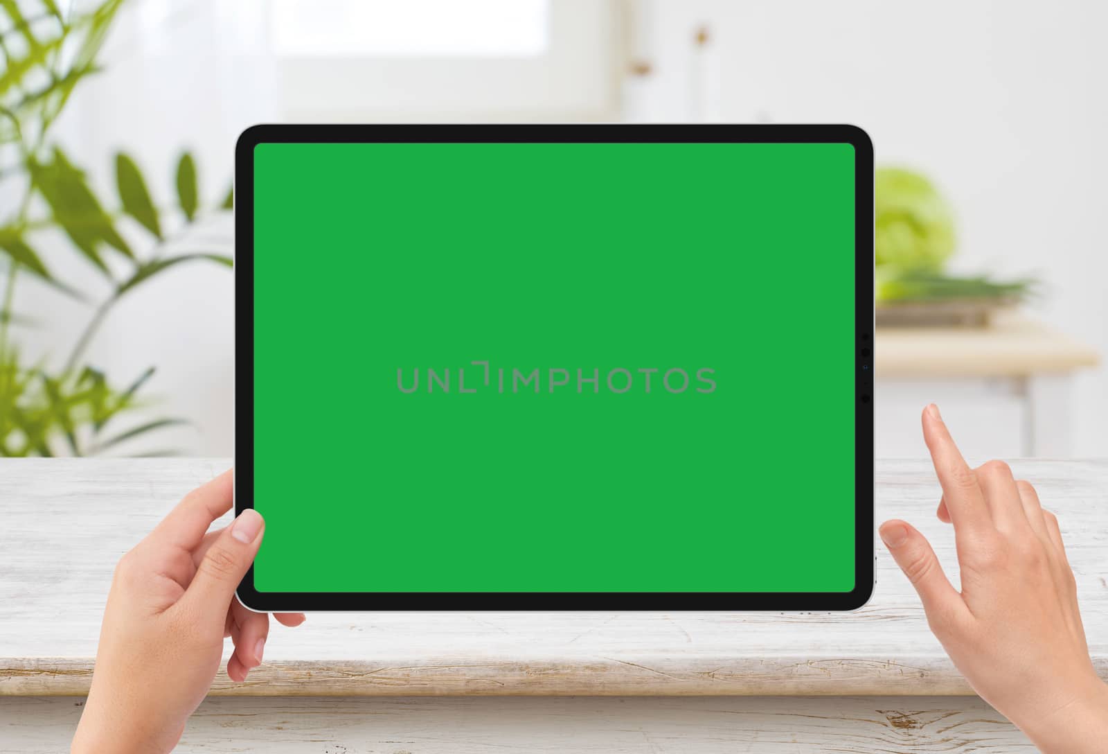 Isolated human left hand holding black tablet media device with white green screen mockup and wooden table in kitchen by cougarsan