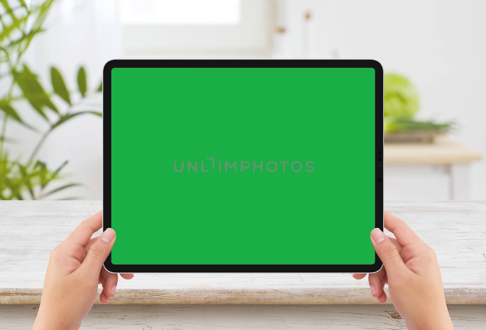 Isolated human two hands holding black tablet media device with white green screen mockup and wooden table in kitchen