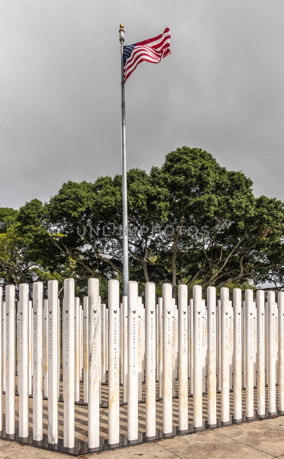 Oahu, Hawaii, USA. - January 10, 2020: Pearl Harbor, Closeup of group of white name-sticks and USA flag at corner on USS Oklahoma memorial. Green foliage in back. Dark cloudscape.