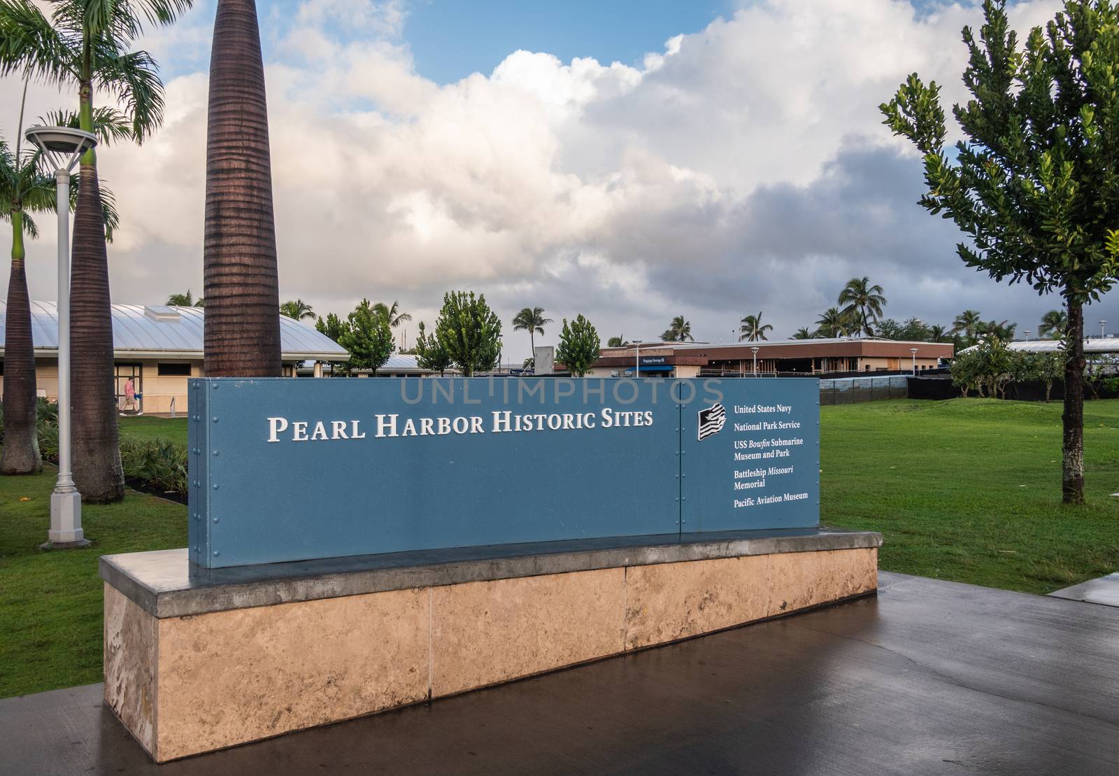 Oahu, Hawaii, USA. - January 10, 2020: Pearl Harbor Historic Sites welcome sign has white letters on blue background under white cloudscape with green lawn in back. Some low level buildings.