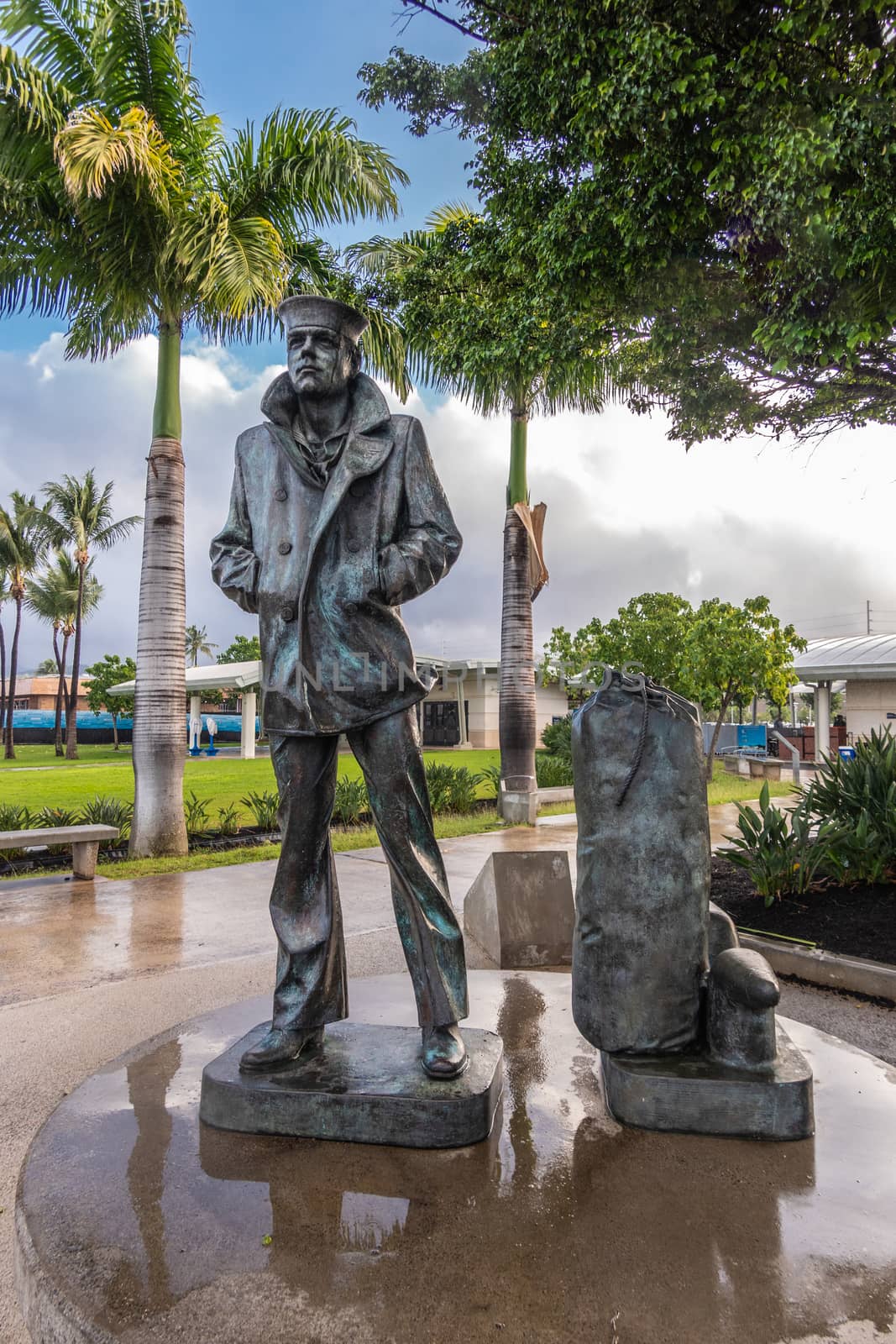 Oahu, Hawaii, USA. - January 10, 2020: Pearl Harbor. Wet Lone waiting sailor bronze statue under green foliage and blue cloudscape. Wet surface around and green lawn in back.