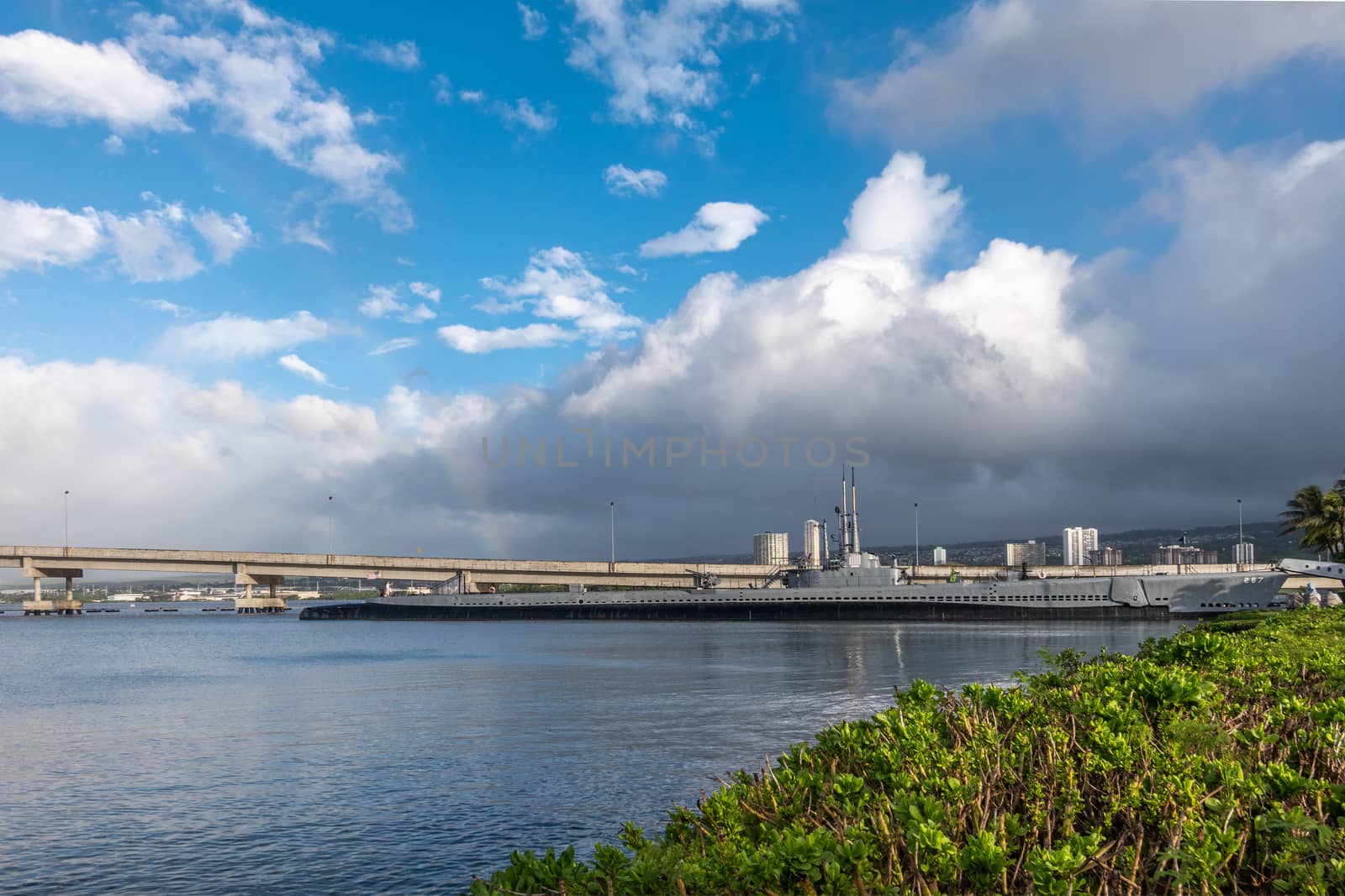 Oahu, Hawaii, USA. - January 10, 2020: Pearl Harbor. Wide shot shows long submarine USS Bowfin sticking out of gray water under blue cloudscape. Ford Island bridge lines up with boat. Green foliage up front.