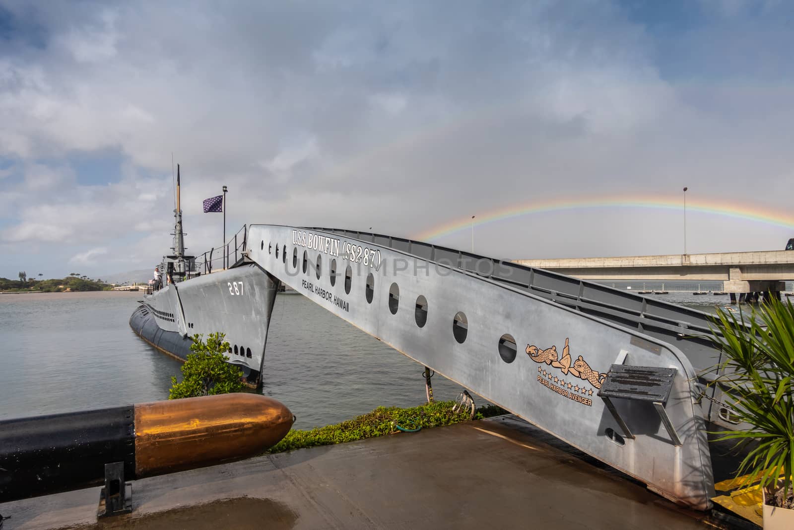 Oahu, Hawaii, USA. - January 10, 2020: Pearl Harbor. Gangway leads up to long submarine USS Bowfin sticking out of gray water under blue cloudscape. Rainbow in sky.