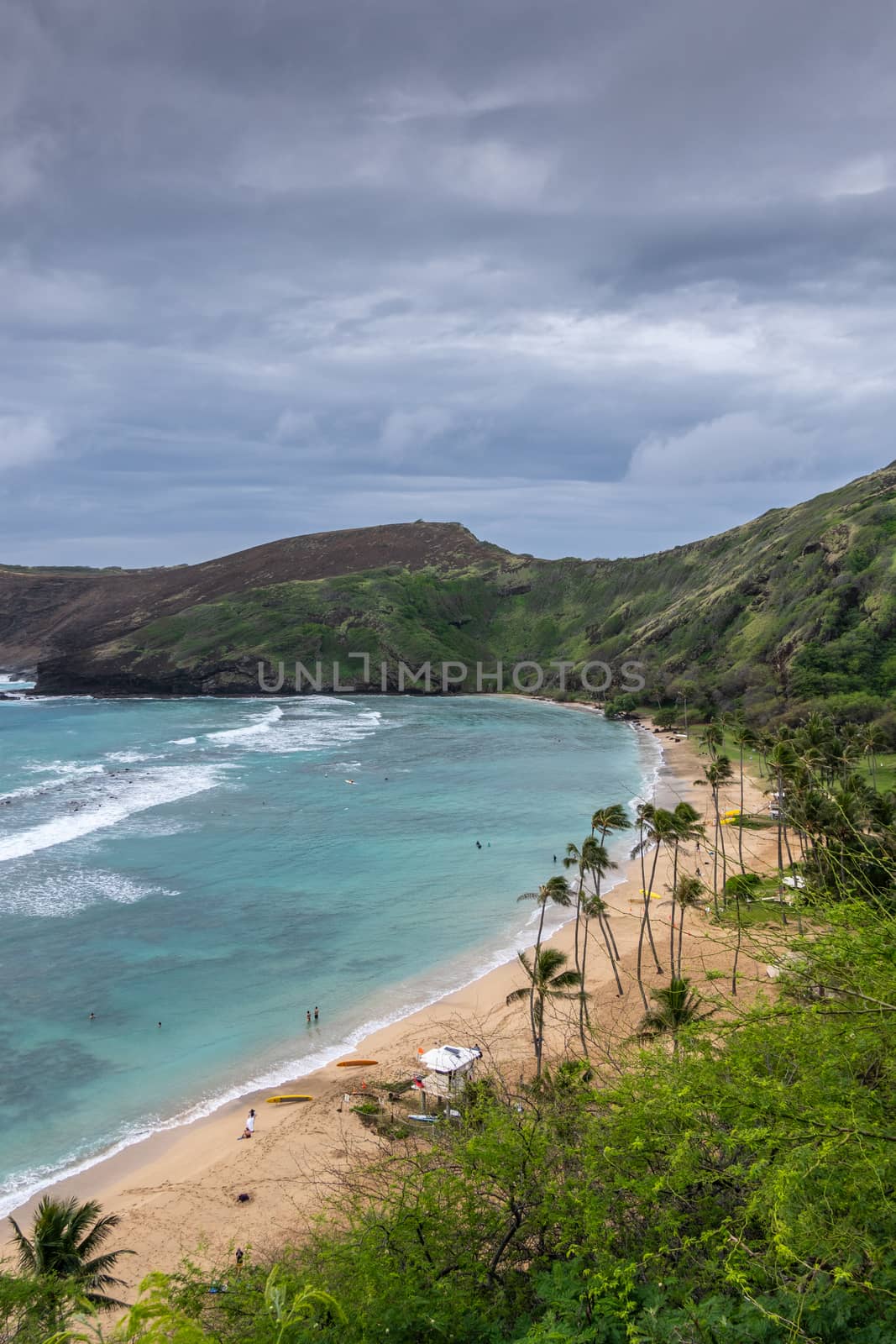 Oahu, Hawaii, USA. - January 11, 2020: Hanauma Bay Nature Preserve. People on Brown sandy beach with palmtrees, white surf on azure water, green cliffs and rocks. All under heavy storm cloudscape.