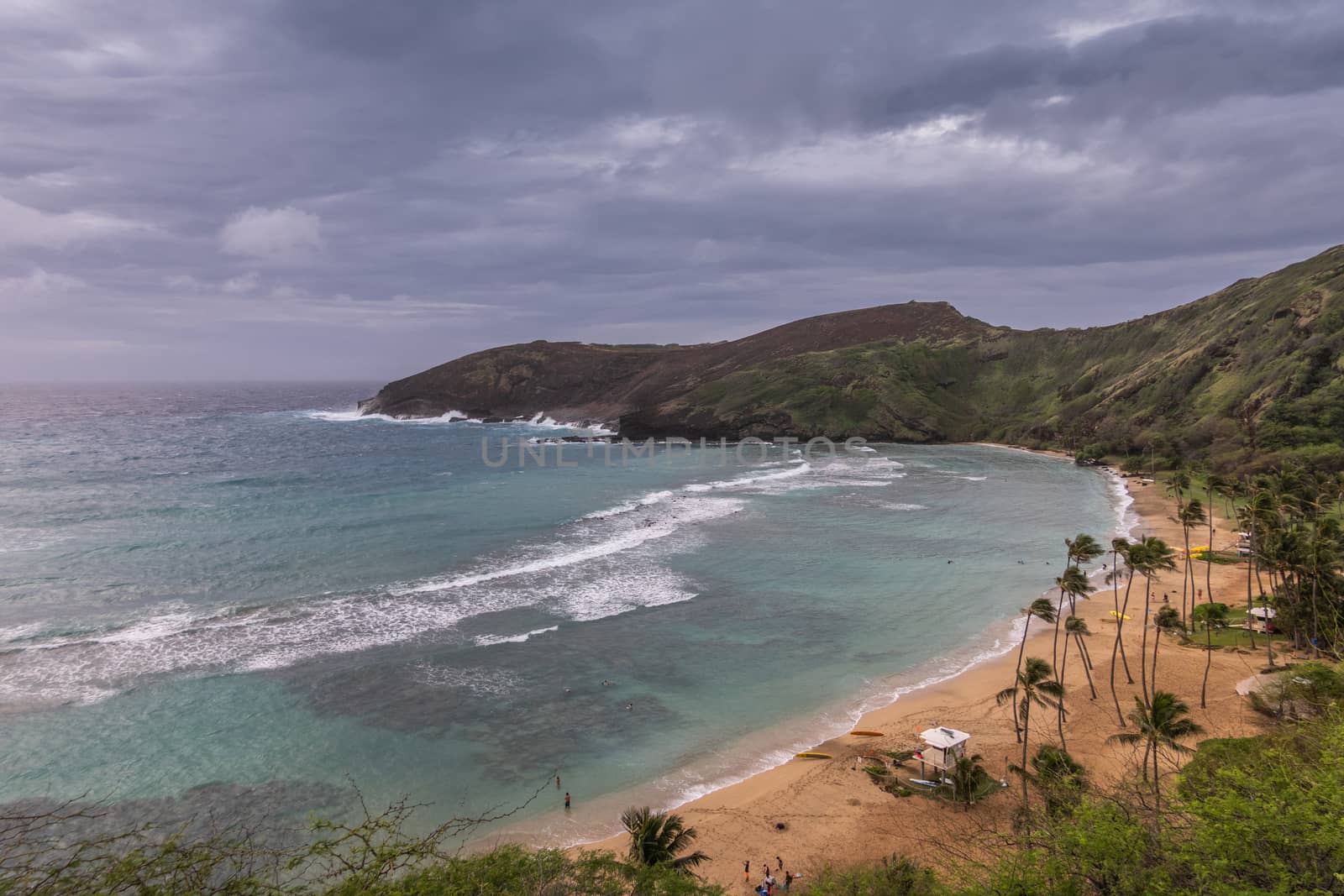 Oahu, Hawaii, USA. - January 11, 2020: Hanauma Bay Nature Preserve. Brown sandy beach with palmtrees, white surf on azure water, dark cliffs and rocks. All under heavy storm cloudscape.