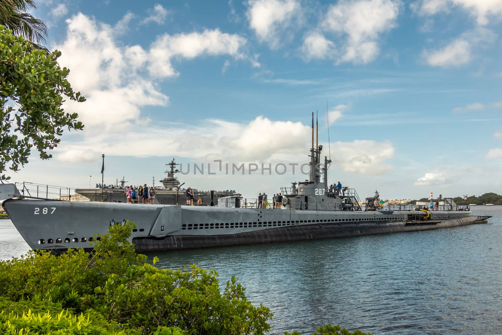 Oahu, Hawaii, USA. - January 10, 2020: Pearl Harbor. Long submarine USS Bowfin sticking out of blueish water under blue cloudscape. People on top. Deck of Abraham Lincoln aircraft carrier in back.