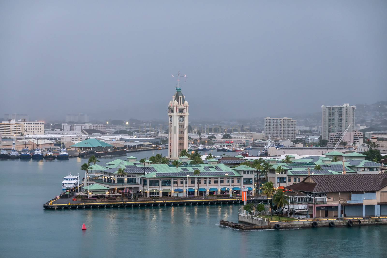 Honolulu  Oahu, Hawaii, USA. - January 11, 2020: white Aloha tower and shopping mall with green roof under dark rainy sky. Wider view on several piers and cityscape.