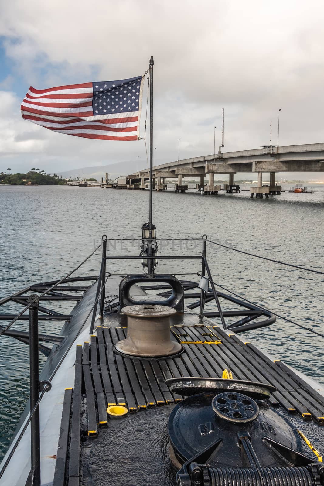 Oahu, Hawaii, USA. - January 10, 2020: Pearl Harbor. Stern with American flag of long submarine USS Bowfin. Part of Ford Island bridge along. All under gray sky with blue patch.