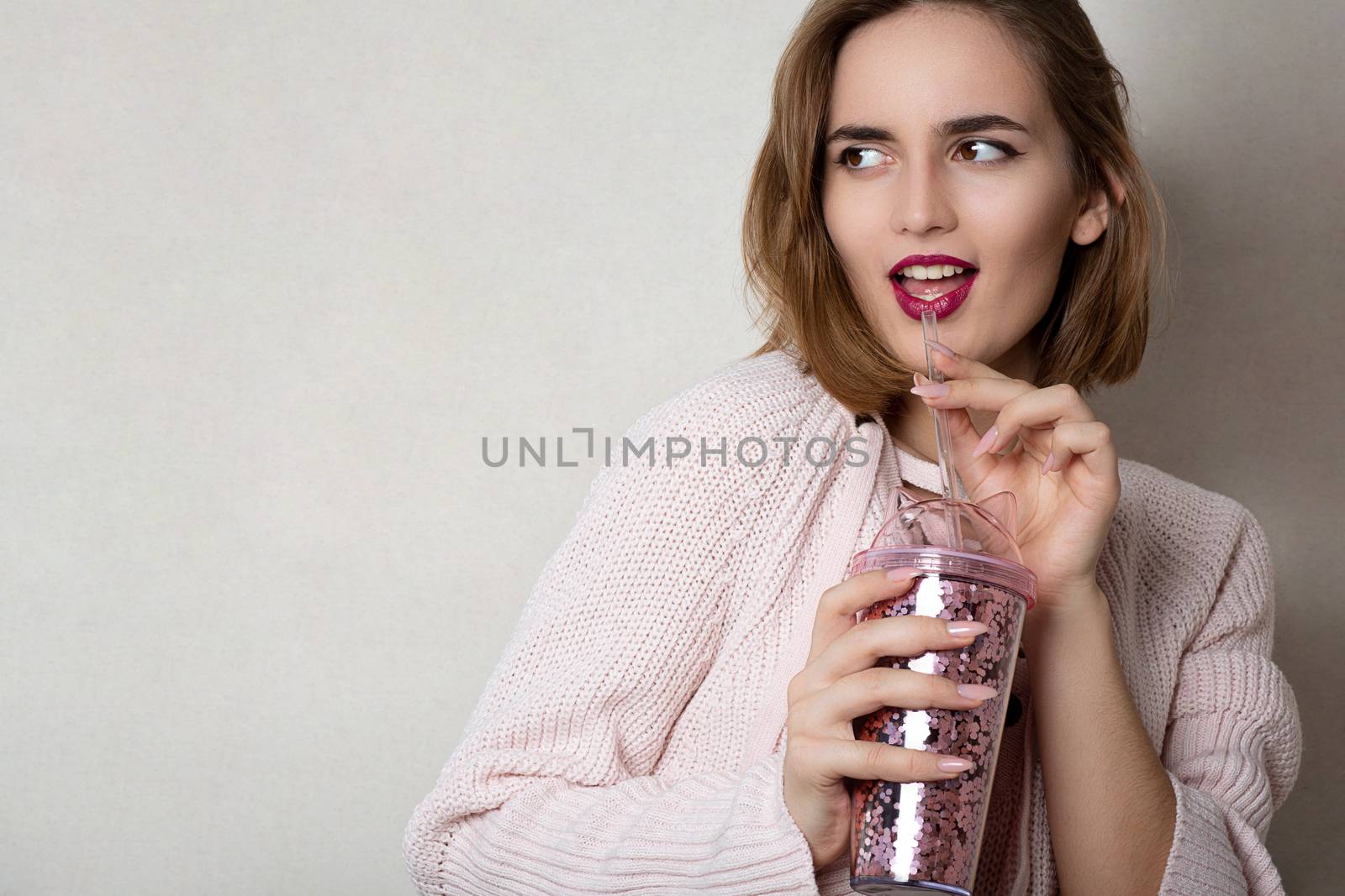 Fashionable young woman wears pink sweater holding glass of orange juice over a grey wall. Space for text