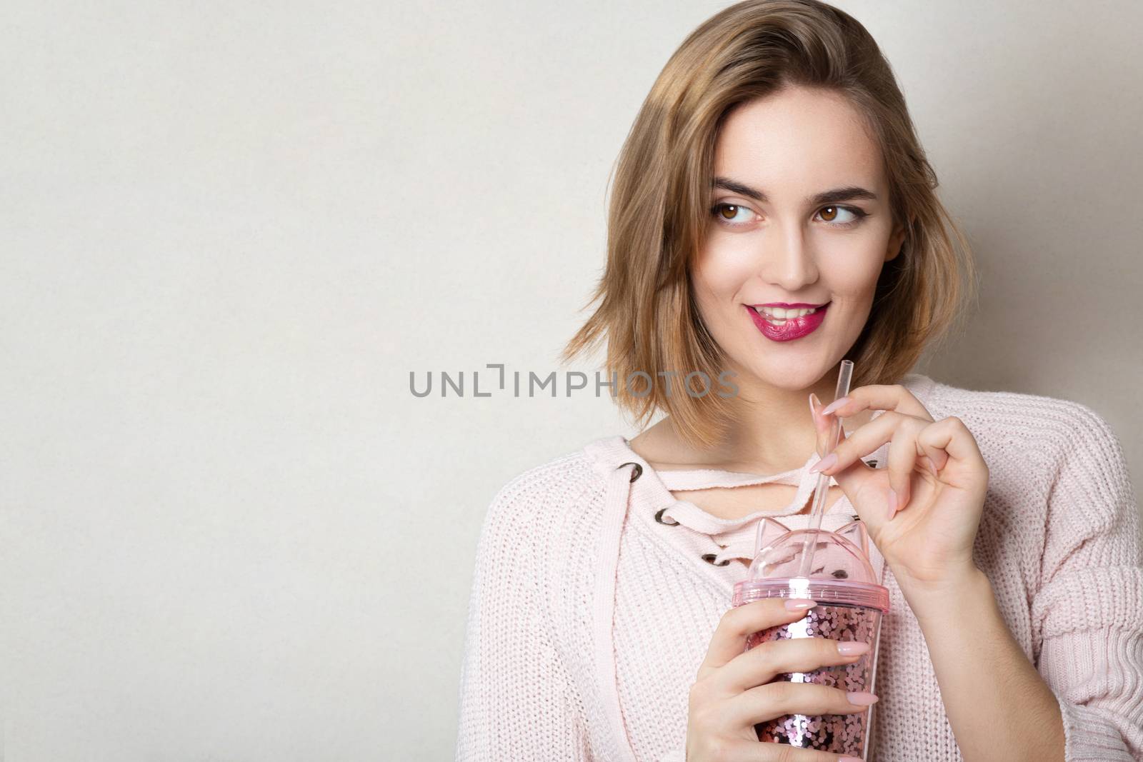 Seductive young woman wears pink sweater holding glass of orange juice over a grey wall. Space for text