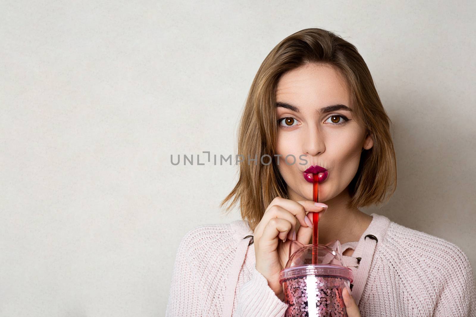 Flirty young woman wears pink sweater holding glass of orange juice over a grey wall. Space for text