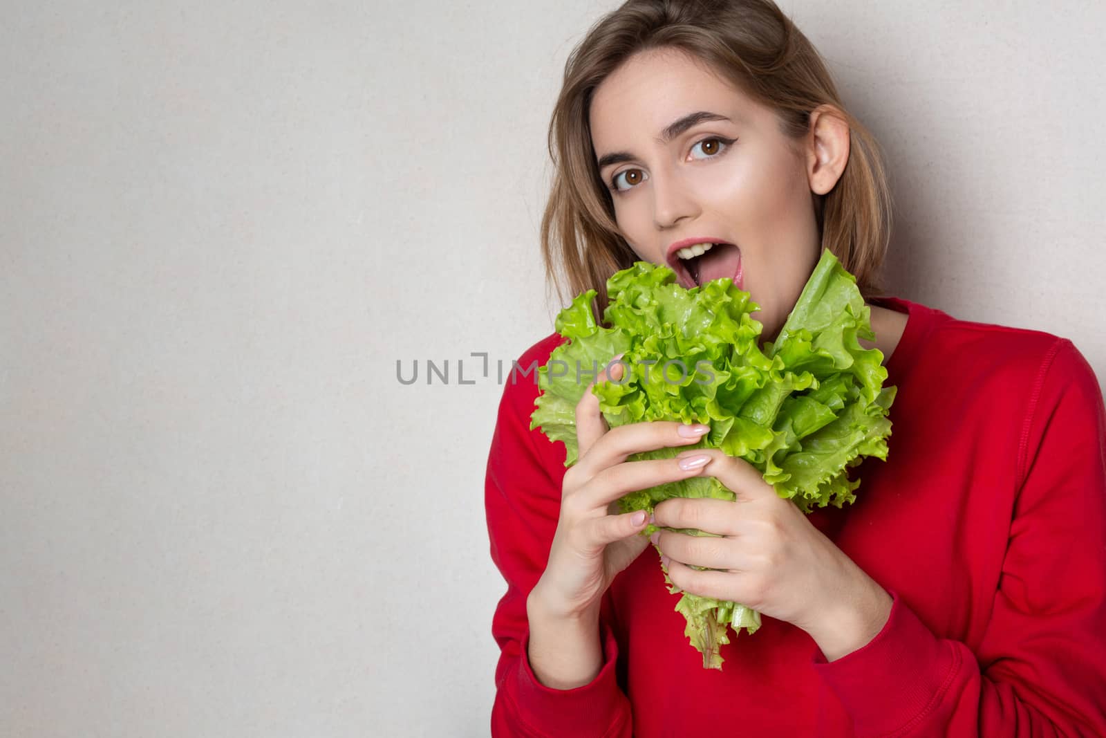 Surprised pretty woman wears red sweater holding lettuce over a grey background. Empty space