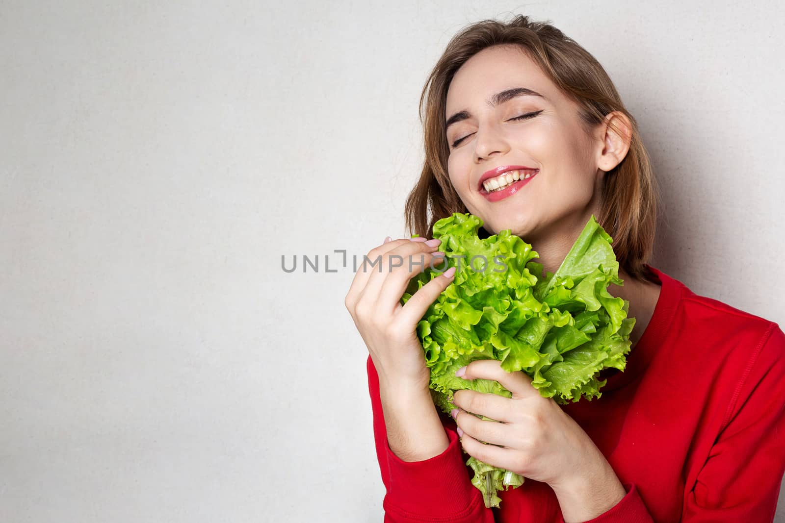 Happy brunette woman wears red sweater holding lettuce over a grey background. Empty space