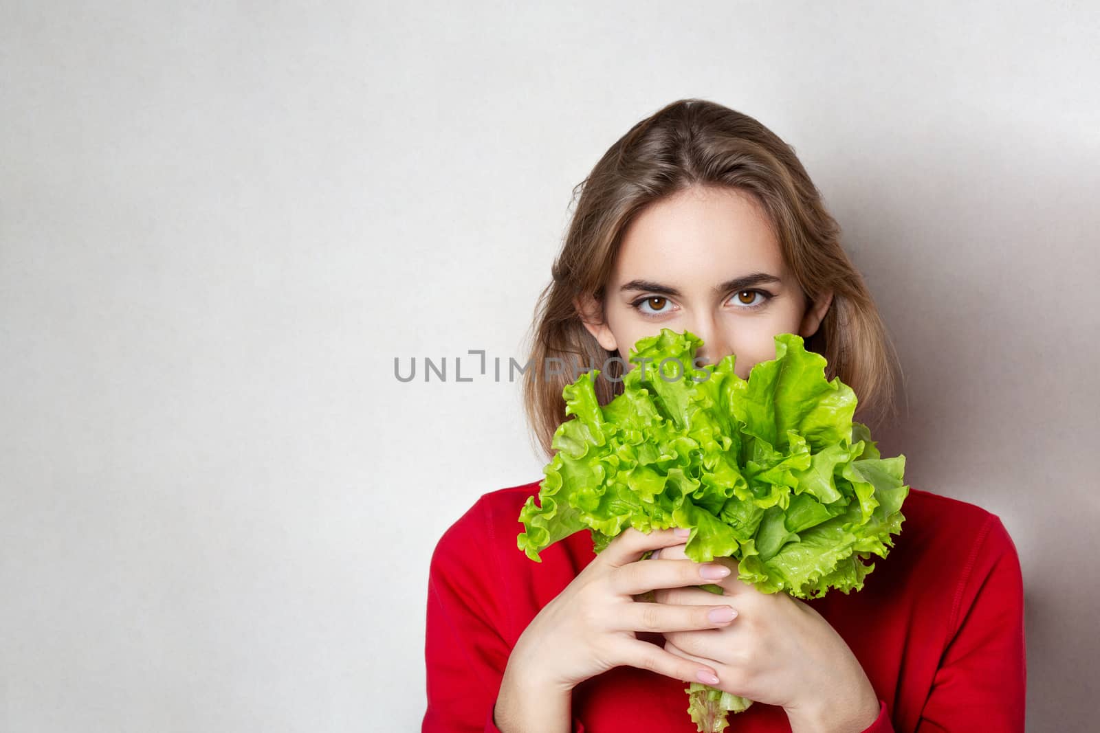 Adorable brunette woman wears red sweater holding lettuce over a grey background. Empty space