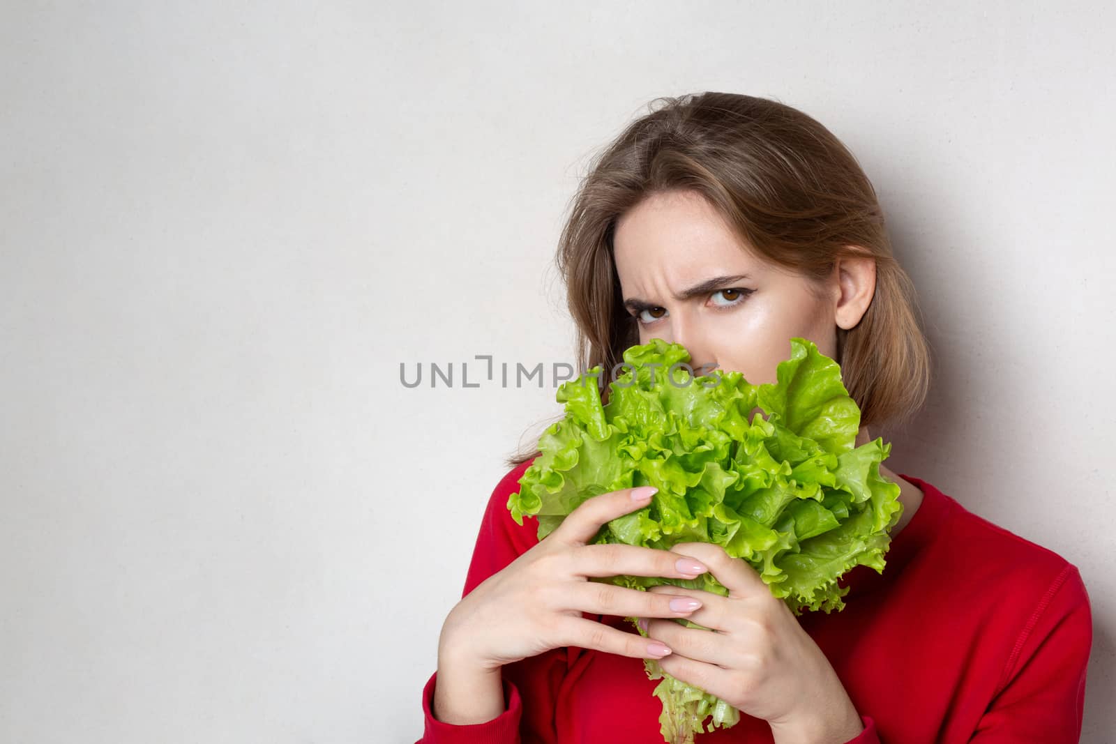 Suspecting brunette woman wears red sweater holding lettuce over a grey background. Empty space