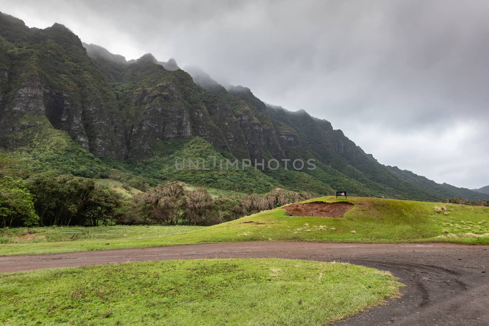 Kaaawa, Oahu, Hawaii, USA. - January 11, 2020: Dirt road makes U-turn ingreen meadow with dark brown to black high rocky cliffs on side of Kualoa valley under cloudy rainy sky.