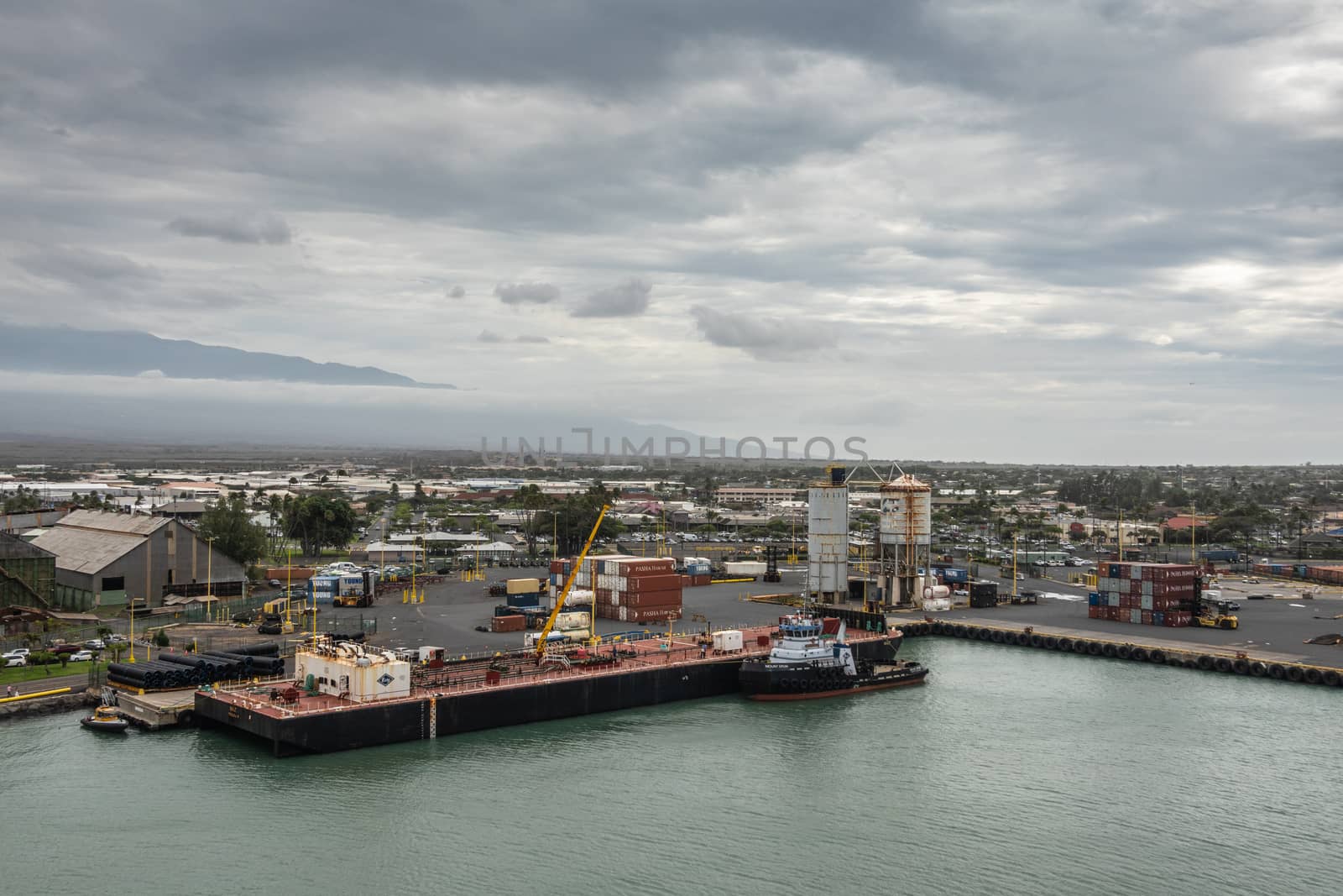 Kahului, Maui,, Hawaii, USA. - January 13, 2020: Nale double hull liquid tank barge held by 1 tugboat against container quay in port under rainy cloudscape. Buildings of town in back.