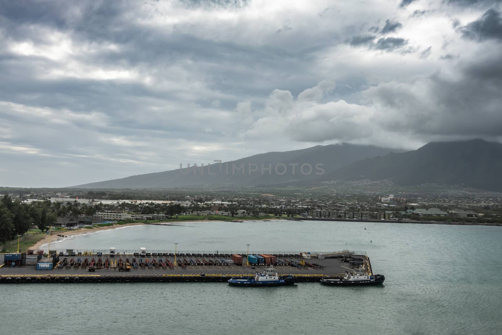 Kahului, Maui,, Hawaii, USA. - January 13, 2020: 2 black mountains tower over cityscape and pier full of shipping container trailers under heavy thick cloudscape full of rain. Tugboars along quay.