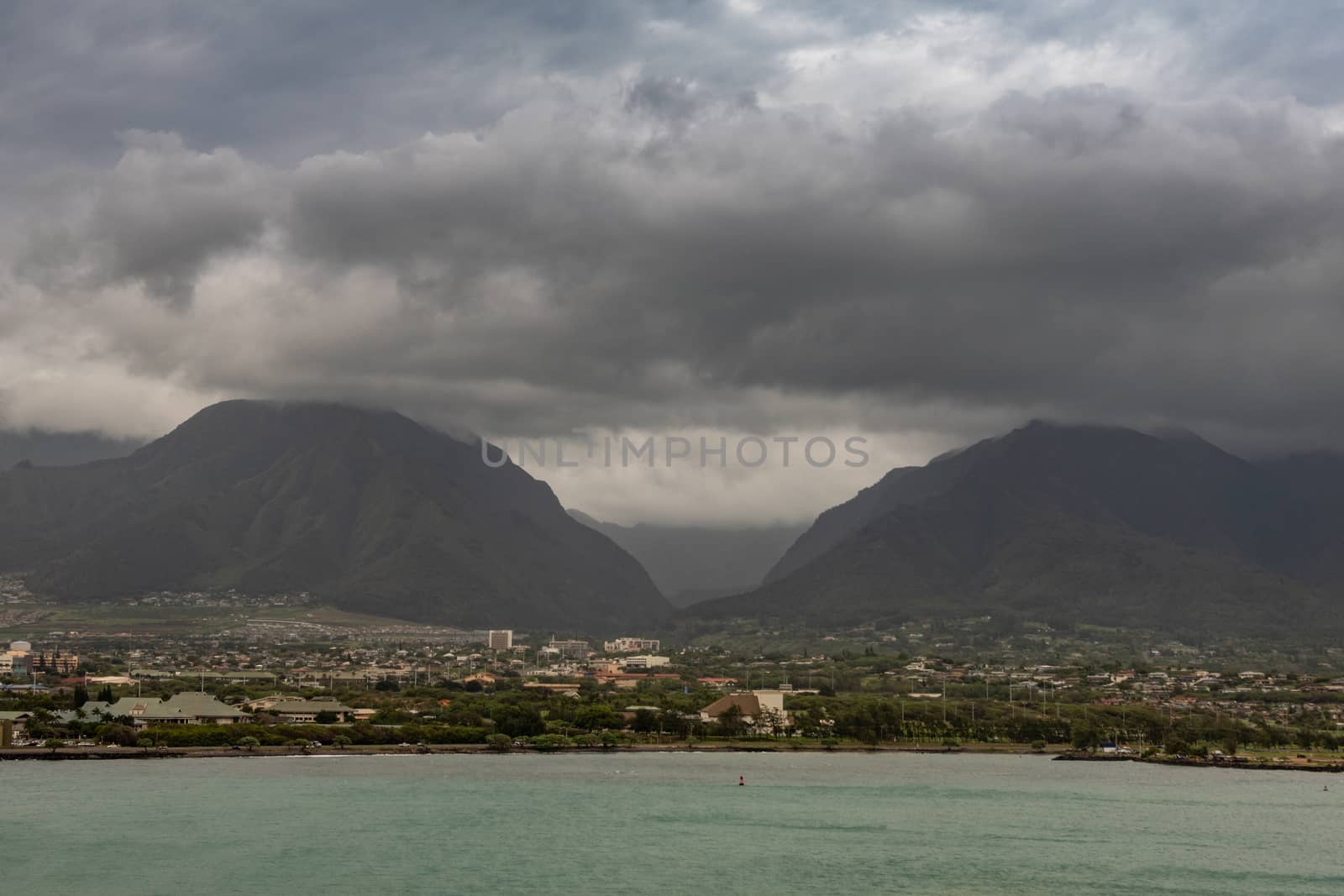 Kahului, Maui,, Hawaii, USA. - January 13, 2020: 2 black mountains tower over cityscape under heavy thick cloudscape full of rain. Azure ocean up front.