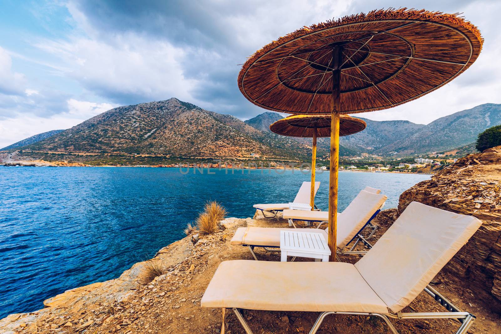 Summer vacation destination. Straw sunshades and sunbeds on the empty rocky beach with sea in the background. Vacation And Tourism Concept. Sunbeds On The Paradise Beach. Umbrellas and sunbeds.