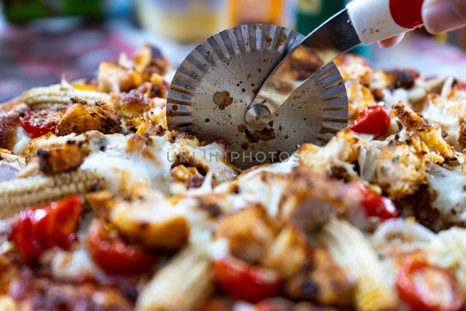 Macro view of a circular pizza cutter curring through a pizza filled with cheese, baby corn, tomatoes, chillies and more on a vegetarian pizza by Shalinimathur