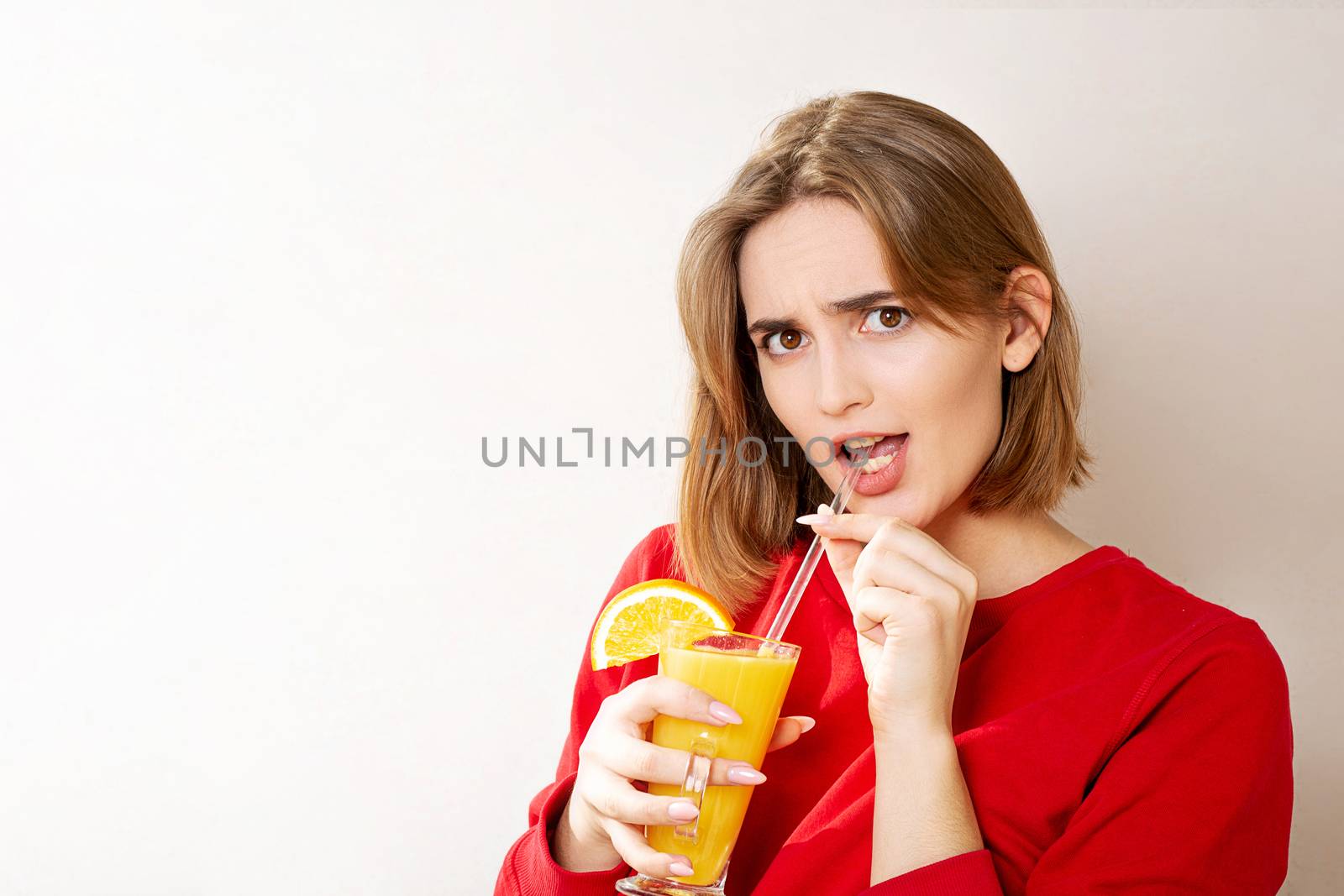 Disappointed young woman wears red sweater holding glass of orange juice over a grey wall. Space for text