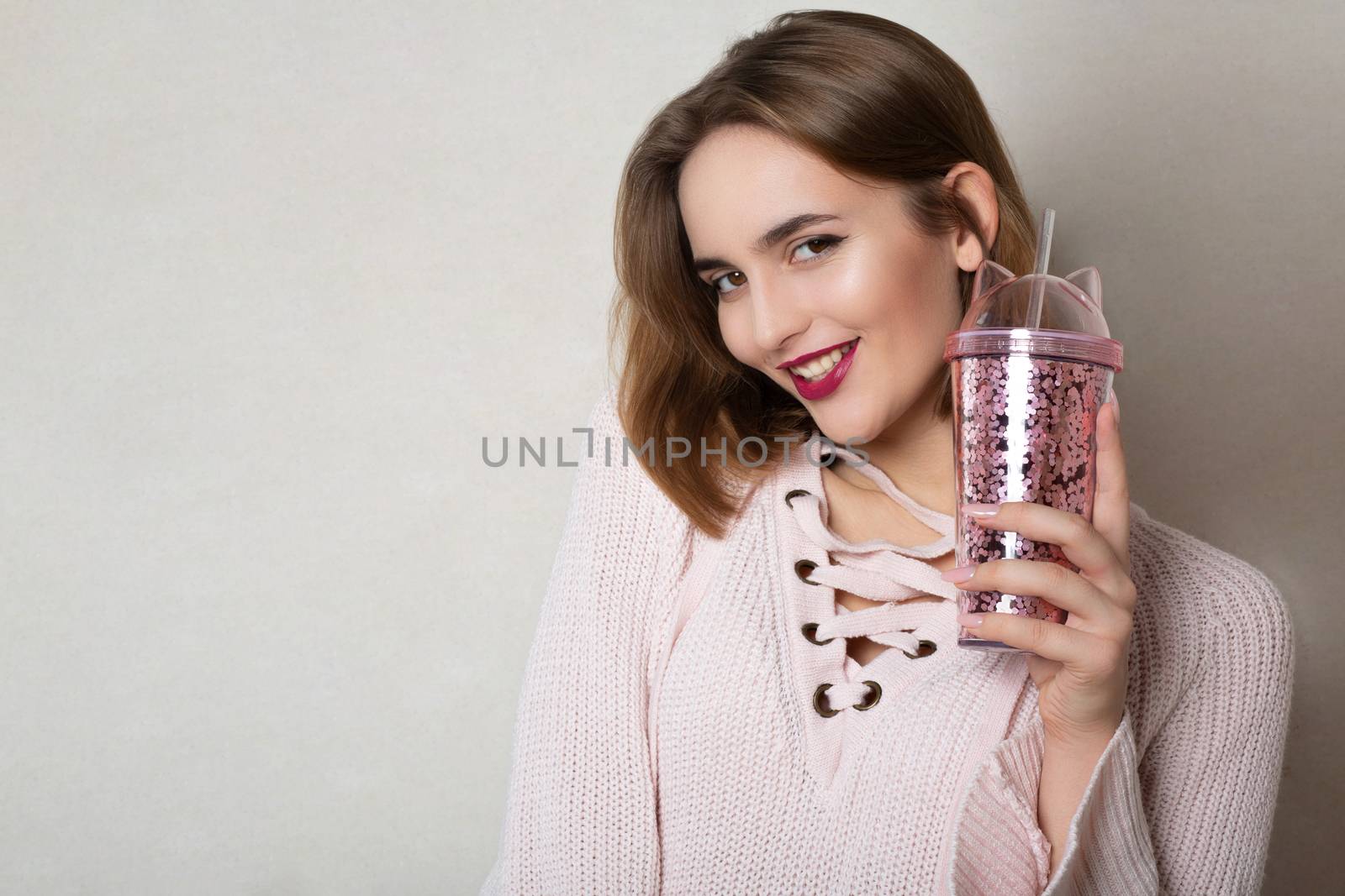 Cheerful young woman wears pink sweater holding glass of orange juice over a grey wall. Space for text