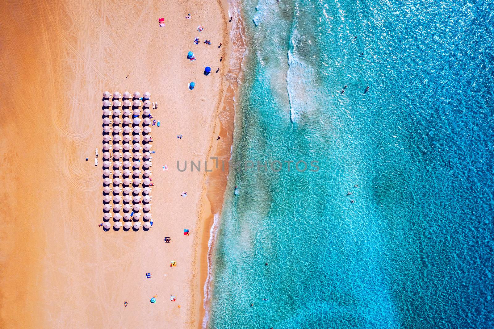 Aerial view of sandy beach with colorful umbrellas, swimming people in sea bay with transparent blue water at sunset in summer. Aerial top view on the beach, umbrellas, sand and sea waves.