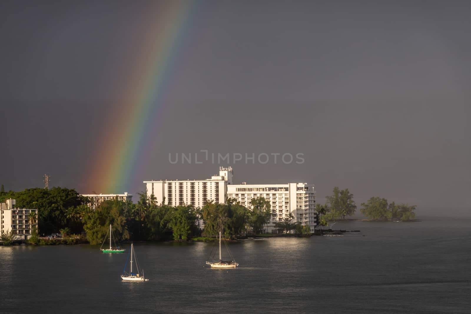 Hilo, Hawaii, USA. - January 14, 2020: Rainbow in dark rain and storm sky touches Grand Naniloa Hotel on shore. Sun on white walls and sides of 3 sailing boats. Green foliage.