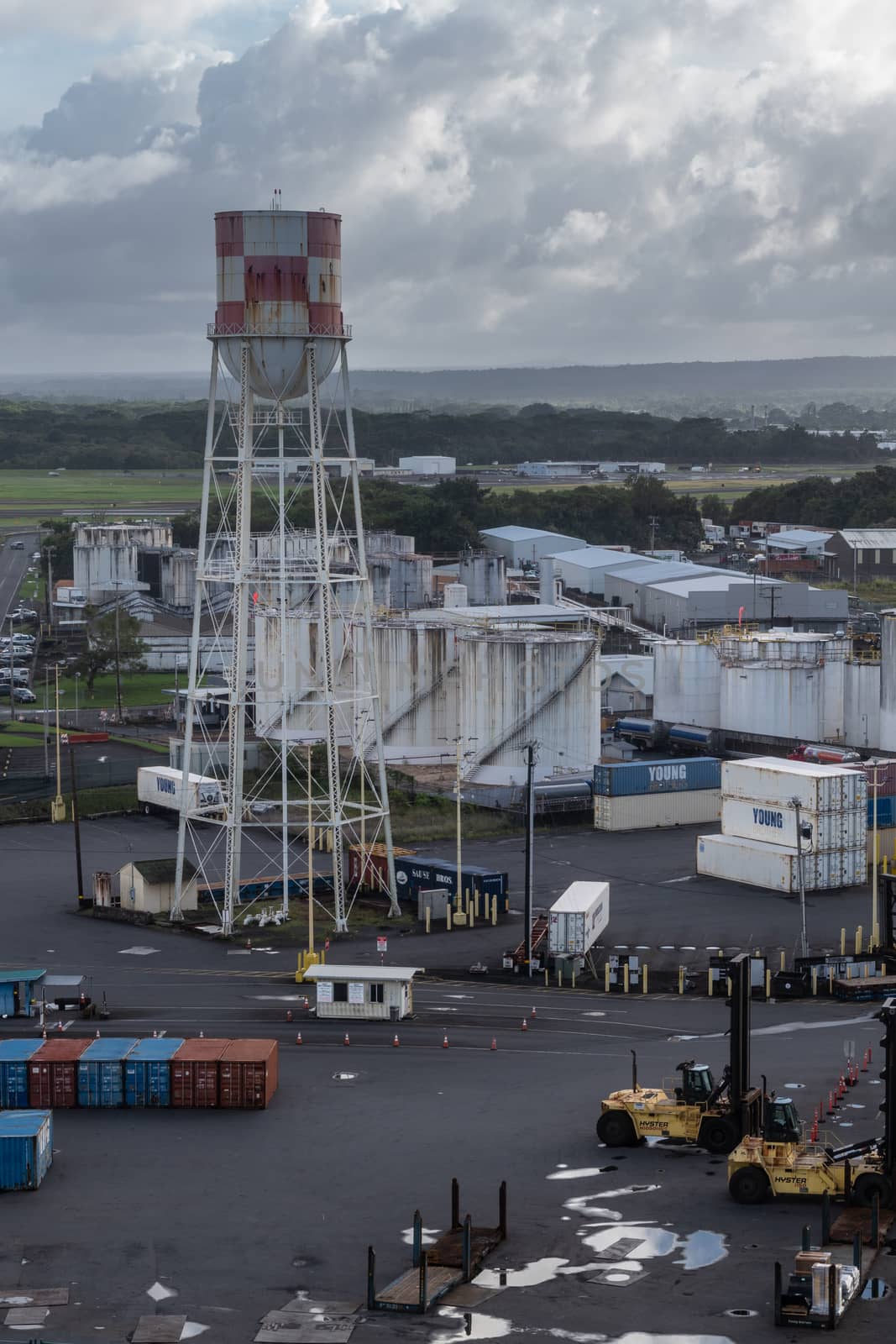 Hilo, Hawaii, USA. - January 14, 2020: Closeup of white and red water tower in port, with fuel tanks in back, shipping containers on the side and crane trucks in front under heavy cloudscape. Green on horizon.