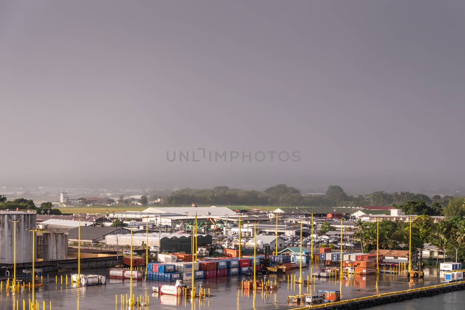 Hilo, Hawaii, USA. - January 14, 2020: Quay with shipping containers in port. Yellow pillars split terrain. Green Airfield in back. Raining with dark sky. Green trees on horizon.