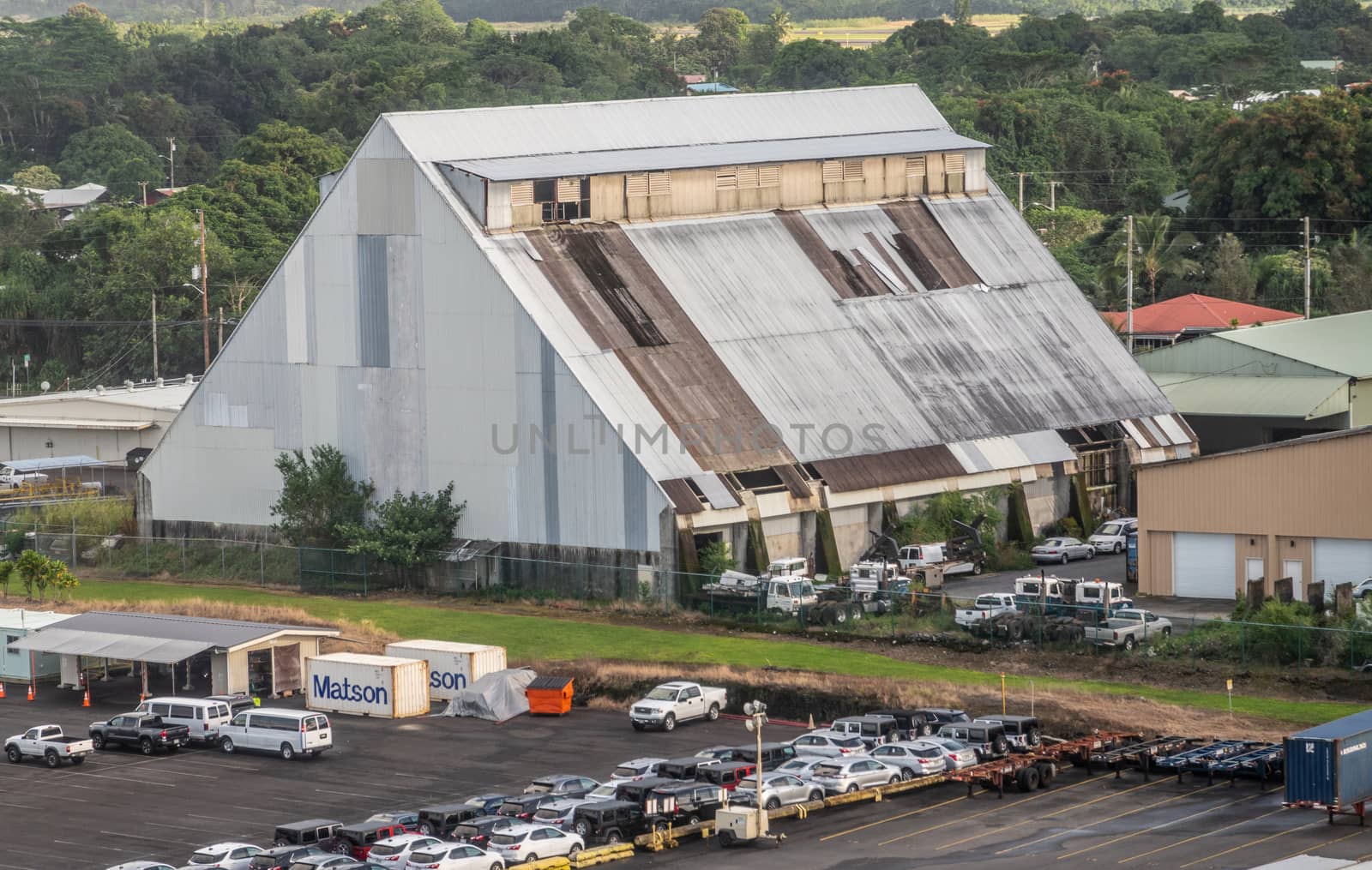 Hilo, Hawaii, USA. - January 14, 2020: Ocean port. Tal gray corrugated plates barn and warehouse with lots of cars and trucks around and green trees and meadow in back.