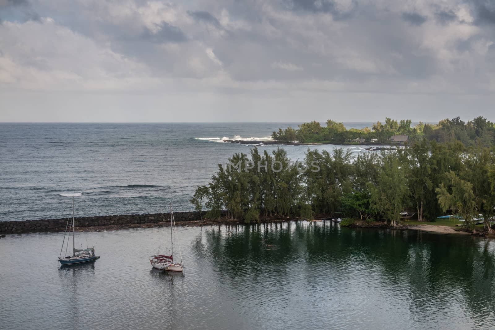 Hilo, Hawaii, USA. - January 14, 2020: Ocean port separated by breakwater from gray-blue ocean under cloudscape. Green trees on shore, some standing in water.